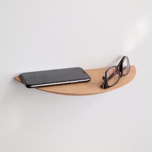 May include: A half-moon shaped wooden shelf mounted on a white wall. A black smartphone and a pair of black-framed eyeglasses rest on the shelf. The shelf is made of light-colored wood.