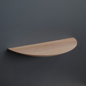 May include: A light-colored, semi-circular wooden shelf mounted on a dark gray wall. The shelf has a smooth, curved edge and a flat surface, showcasing the natural wood grain. It is designed for displaying small items.