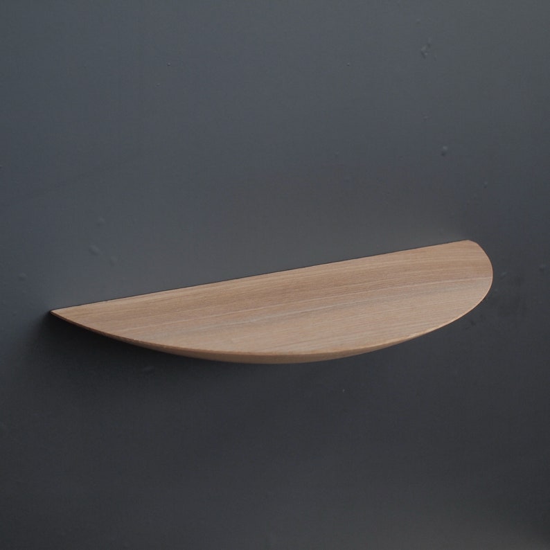 May include: A light-colored, semi-circular wooden shelf mounted on a dark gray wall. The shelf has a smooth, curved edge and a flat surface, showcasing the natural wood grain. It is designed for displaying small items.