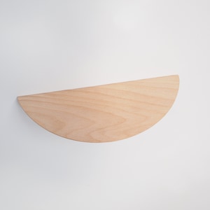May include: A half-circle wooden shelf, crafted from light-colored wood, is mounted on a white wall. The shelf's smooth surface and curved edge create a minimalist design. This floating shelf is ideal for displaying small items or adding a touch of modern decor.