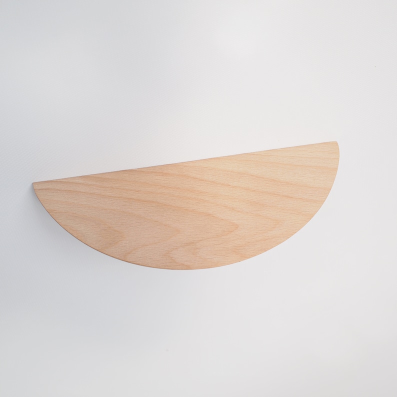 May include: A half-circle wooden shelf, crafted from light-colored wood, is mounted on a white wall. The shelf's smooth surface and curved edge create a minimalist design. This floating shelf is ideal for displaying small items or adding a touch of modern decor.