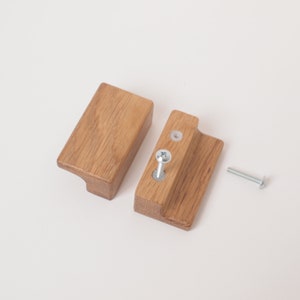 May include: Two wooden cabinet knobs with a rectangular shape and a slight curve on the bottom. The knobs are made of light brown wood and have a silver screw in the top.