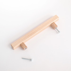 May include: A light wood cabinet pull with a rectangular shape and rounded edges. The pull is shown with two screws.