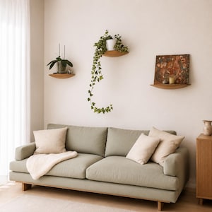 May include: A light green sofa with cream-colored pillows and a white blanket. Wooden floating shelves display potted plants and artwork. The room has a minimalist, modern aesthetic with natural light.