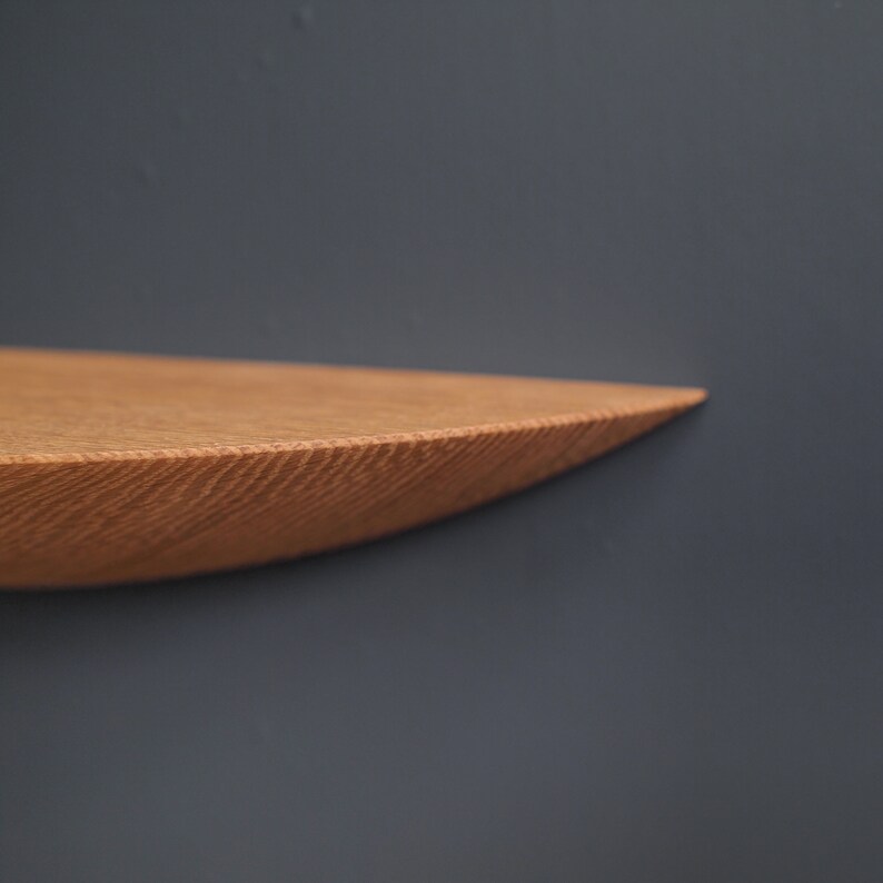 K&ouml;nnte beinhalten: Nahaufnahme einer Holzregals mit abgerundeter Kante. Das Regal ist aus hellbraunem Holz und hat eine nat&uuml;rliche Maserung. Das Regal ist an einer dunkelgrauen Wand montiert.
