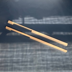 Könnte beinhalten: Zwei rechteckige Holzknaufgriffe für Schranktüren mit einem ausgeschnittenen Griff. Die Griffe sind aus hellem braunem Holz und liegen auf einer dunkelgrauen Oberfläche.