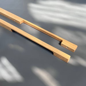 May include: Two wooden cabinet pulls with a rectangular shape and a slight curve at the top. The pulls are made of light brown wood and have a natural finish.