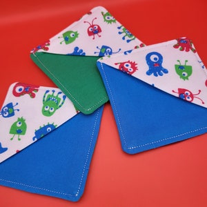 May include: Three colorful fabric bookmarks with a white background and a pattern of cartoon monsters in red, blue, and green. The bookmarks are folded in half and have a blue, green, and blue fabric on the back.
