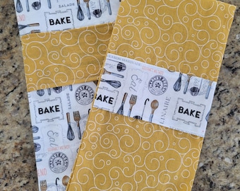 May include: Two kitchen towels with a yellow and white pattern. The towels have a white band with the words "Bake" and "Market Fresh" printed on them. The towels are folded and stacked on top of each other.