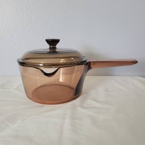 May include: A vintage amber-colored glass saucepan with a matching lid. The saucepan has a single handle and a pouring spout. The lid has a small, dark brown knob. The glass is transparent, allowing the contents to be seen. A classic kitchenware item.