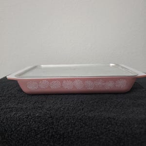 May include: A rectangular, pink Pyrex baking dish with a clear glass lid. The dish features a white daisy pattern along the sides. The dish is sitting on a black surface.