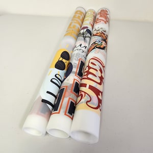 Puede incluir: Cuatro impresiones decorativas enrolladas con varios dise&ntilde;os. Una presenta gafas de sol, otra tiene elementos florales y de texto, y las otras muestran texto y gr&aacute;ficos en negrita en naranja y rojo. Las impresiones son blancas con dise&ntilde;os coloridos.