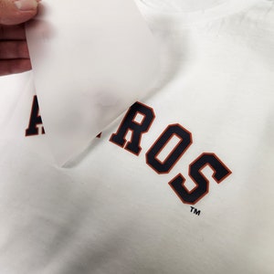 Puede incluir: Camiseta blanca con la palabra "ASTROS" impresa en azul marino con ribete naranja. Una mano despega una l&aacute;mina transl&uacute;cida de la camiseta, revelando el dise&ntilde;o. La camiseta tiene un cuello redondo cl&aacute;sico.