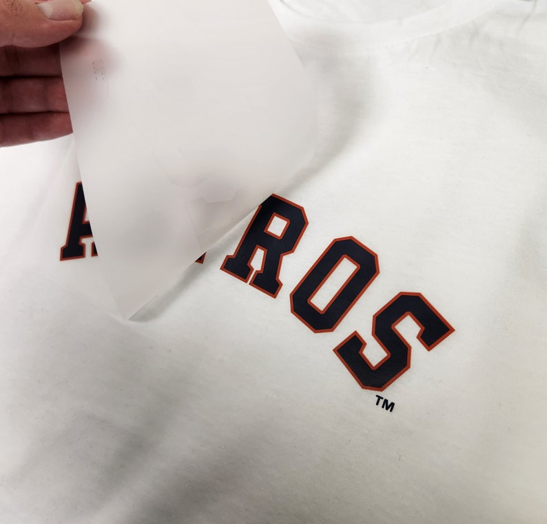 Puede incluir: Camiseta blanca con la palabra "ASTROS" impresa en azul marino con ribete naranja. Una mano despega una l&aacute;mina transl&uacute;cida de la camiseta, revelando el dise&ntilde;o. La camiseta tiene un cuello redondo cl&aacute;sico.