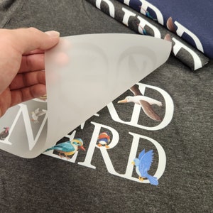 Puede incluir: Una camiseta gris con la palabra "NERD" en letras blancas, con ilustraciones de p&aacute;jaros coloridos. Una mano levanta una l&aacute;mina transl&uacute;cida que revela el dise&ntilde;o. Se ven camisetas adicionales con dise&ntilde;os de p&aacute;jaros.