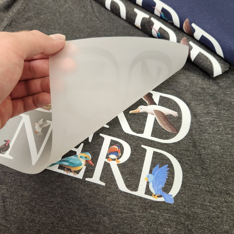 Puede incluir: Una camiseta gris con la palabra "NERD" en letras blancas, con ilustraciones de p&aacute;jaros coloridos. Una mano levanta una l&aacute;mina transl&uacute;cida que revela el dise&ntilde;o. Se ven camisetas adicionales con dise&ntilde;os de p&aacute;jaros.