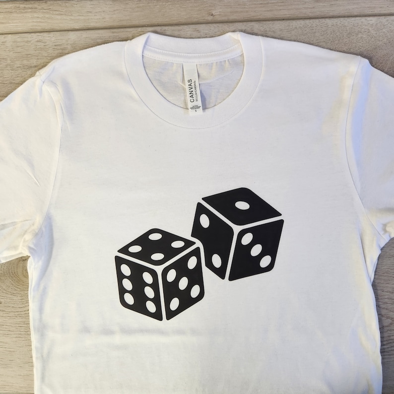 Puede incluir: Camiseta blanca con un gr&aacute;fico negro de dos dados. Los dados se muestran con sus patrones de puntos, uno mostrando un seis y el otro un uno. La camiseta est&aacute; hecha de un material suave.