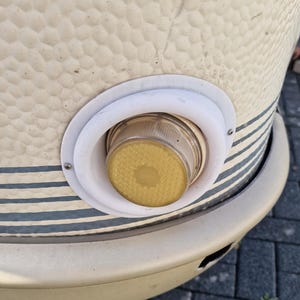 Op de afbeelding: Een crèmekleurig, getextureerd, cilindrisch object met een cirkelvormig, geel getint lichtarmatuur in een witte kunststof behuizing. Het buitenoppervlak vertoont een herhalend noppenpatroon en horizontale donkergrijze strepen.
