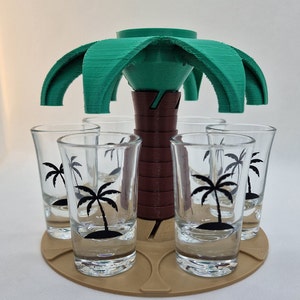 Könnte beinhalten: Ein grüner, palmenförmiger Schnapsglas-Spender mit sechs Schnapsgläsern auf einem braunen Sockel. Die Schnapsgläser haben Palmenmotive.