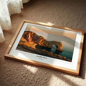 May include: A framed print of Durdle Door in Dorset, England, featuring a natural limestone arch over the sea. The image has warm tones, with the text "Durdle Door Dorset" at the bottom. The frame is light wood.
