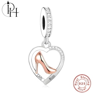 May include: A silver charm with a heart-shaped pendant featuring a rose gold high-heeled shoe. The heart is outlined with clear crystals and has the words "Lady" and "Boss" inscribed. The charm has a silver loop with clear crystals.