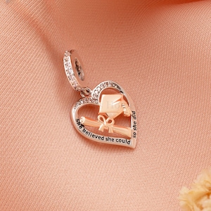 May include: A silver and rose gold charm in the shape of a heart, with a graduation cap and diploma design. The charm has the words "She believed she could so she did" engraved on it. The charm is adorned with clear crystals.