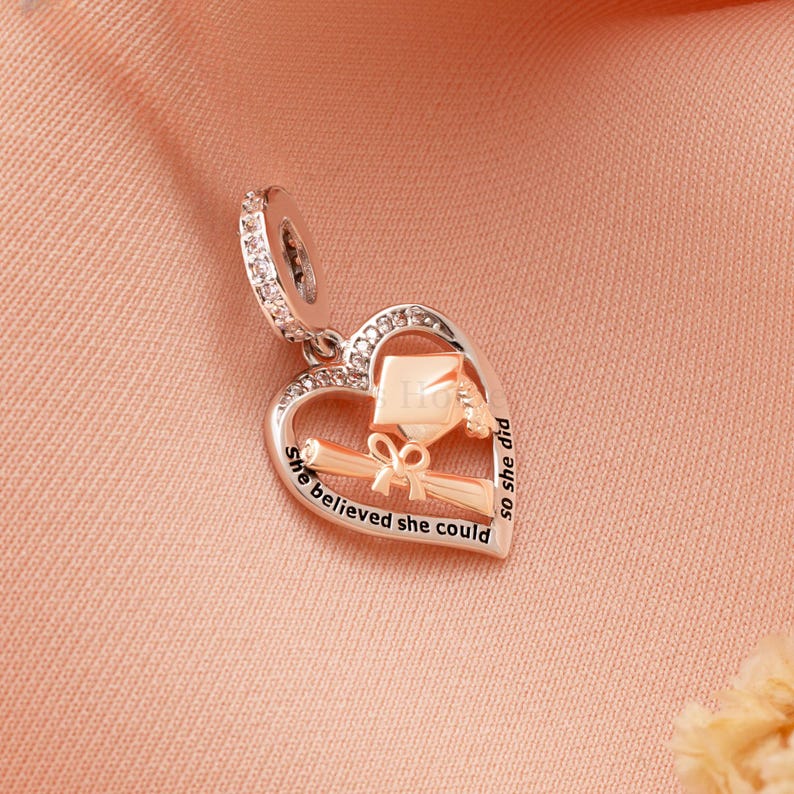 May include: A silver and rose gold charm in the shape of a heart, with a graduation cap and diploma design. The charm has the words "She believed she could so she did" engraved on it. The charm is adorned with clear crystals.