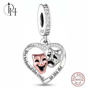 May include: A sterling silver heart-shaped charm featuring a rose gold-toned comedy mask and a silver tragedy mask. The frame is engraved with the text "She believed she could so she did" and accented with small clear stones on the bail and top edge.