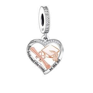 May include: Silver charm with a heart-shaped pendant. The pendant features a graduation cap and diploma in rose gold, with the words "She believed she could so she did" in a cursive font. The charm has a loop with clear crystals.