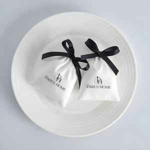 May include: Two small white fabric pouches with black ribbon ties, each bearing the "PARUS HOME" logo in black. The pouches rest in a white, shallow, round dish.