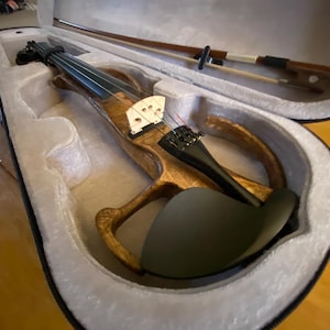 May include: A wooden electric violin with a black fingerboard and a black chin rest rests inside a white lined case. The violin bow is also in the case. The violin has a light brown finish.