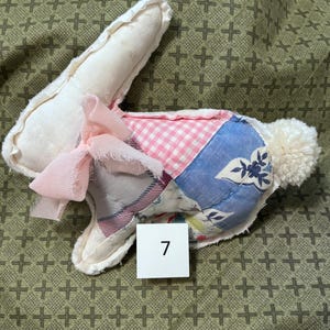 May include: A handmade stuffed bunny with a patchwork design. The bunny features a white head and ears, a pink bow, and a body made of various patterned fabrics, including pink gingham and blue floral prints. A white pom-pom tail completes the design. The number 7 is on a white square.