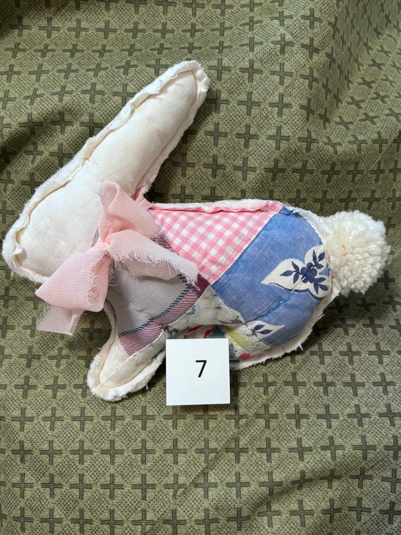 May include: A handmade stuffed bunny with a patchwork design. The bunny features a white head and ears, a pink bow, and a body made of various patterned fabrics, including pink gingham and blue floral prints. A white pom-pom tail completes the design. The number 7 is on a white square.