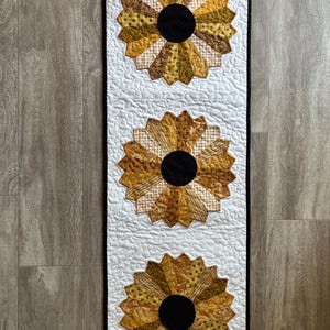 May include: A quilted table runner featuring three large sunflower designs. The sunflowers have a black center and petals in shades of yellow, gold, and brown. The background is white with a quilted pattern, and the edges are trimmed with black fabric.