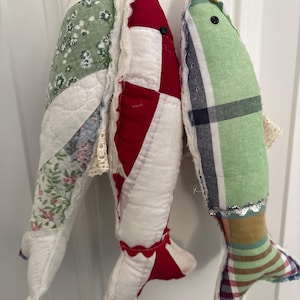 May include: Three stuffed fabric fish ornaments hanging vertically. The left fish features green floral and white quilted patterns. The center fish is red and white. The right fish displays a green plaid pattern with a silver trim accent.
