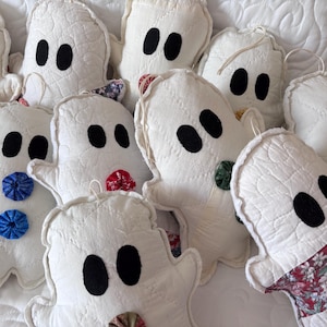 May include: A collection of white fabric ghost ornaments featuring quilted textures and black felt eyes. Each ghost is decorated with small, colorful fabric yo-yo accents or floral patterned patches, and includes a thin string loop for hanging.