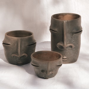 Könnte beinhalten: Drei graue Keramik-Gesichtstöpfe in verschiedenen Größen. Jeder Topf hat ein stilisiertes Gesicht mit geschlossenen Augen und einer markanten Nase. Die Töpfe sind auf einer weißen Oberfläche angeordnet.