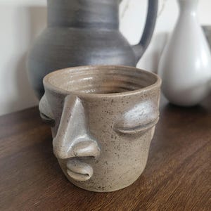 May include: A small, tan ceramic planter shaped like a face with closed eyes and a prominent nose. The planter has a rough, textured surface and is set against a blurred background with other ceramic vases.