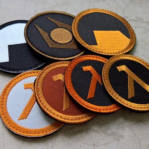Pode incluir: Uma coleção de sete emblemas bordados circulares com vários designs. Alguns emblemas exibem a letra grega lambda em laranja ou dourado sobre fundos pretos, castanhos ou azul-claros, enquanto outros apresentam um logótipo circular estilizado.
