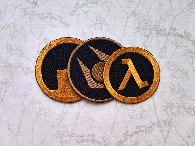 May include: Three circular embroidered patches featuring black and gold designs. The patches display the Half-Life lambda symbol and the Aperture Science logo, each with a thick gold border and detailed stitching on a dark fabric background.