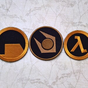 May include: Three circular embroidered patches featuring gold-colored thread designs on a black background. The left patch shows a partial square, the center patch displays a stylized geometric emblem, and the right patch features a Greek lambda symbol.