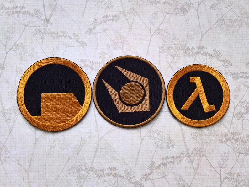 May include: Three circular embroidered patches featuring gold-colored thread designs on a black background. The left patch shows a partial square, the center patch displays a stylized geometric emblem, and the right patch features a Greek lambda symbol.