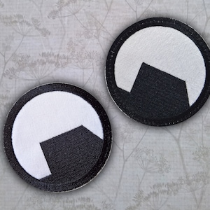 Peut inclure: Deux patchs brodés en noir et blanc avec un motif géométrique. Les patchs sont circulaires et ont une bordure noire.