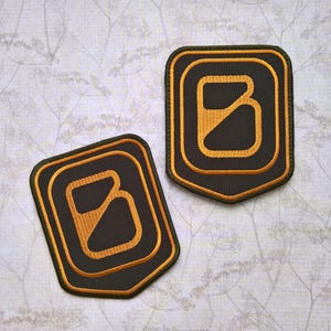 Könnte beinhalten: Zwei gestickte Patches mit schwarzem Hintergrund und goldener Umrandung. Die Patches zeigen einen stilisierten Buchstaben "B" mit einem diagonalen Strich durch ihn.
