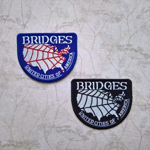 May include: Two embroidered patches with a map of the United States and the text "BRIDGES UNITED CITIES OF AMERICA". The top patch is blue with a red spiderweb design. The bottom patch is black with a white spiderweb design.