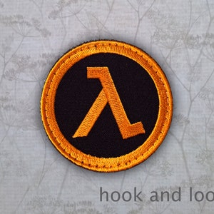 May include: A round, black patch with an orange Lambda symbol in the centre. The patch has a yellow border and is designed for hook and loop attachment.