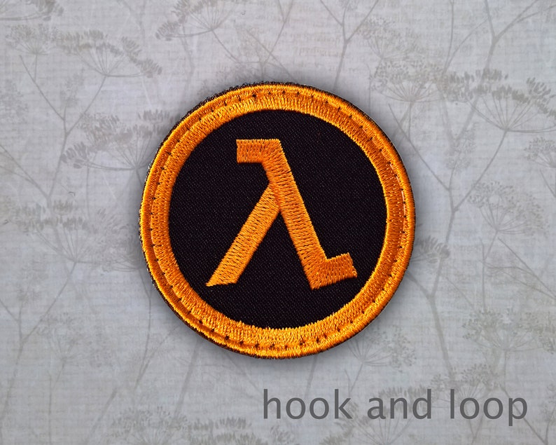 May include: A round, black patch with an orange Lambda symbol in the centre. The patch has a yellow border and is designed for hook and loop attachment.