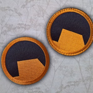Peut inclure: Deux patchs ronds brodés noirs et orange avec un motif géométrique. Les patchs ont un fond noir avec une forme géométrique orange au centre.