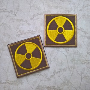 May include: Two brown fabric patches with yellow embroidered radiation symbols. The patches are rectangular and have a beige border.