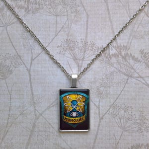 May include: Silver-toned necklace with a rectangular pendant. The pendant features a design with a yellow atom symbol, blue accents, and the word "МОНОЛИТ" in white. The chain is a delicate, linked style.